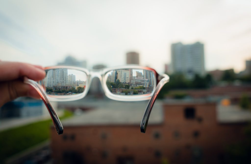 Someone holding up a pair of glasses with the surrounding city background blurred out but sharp focus through the lenses of the glasses to simulate astigmatism