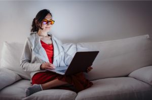 A young woman sitting on a couch with her laptop in her lap, wearing blue light blocking glasses while reading her computer screen