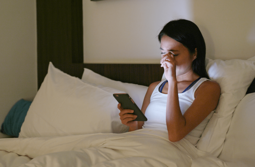 A woman looking at her smartphone before she goes to sleep and she is rubbing her left eye due to dryness and fatigue
