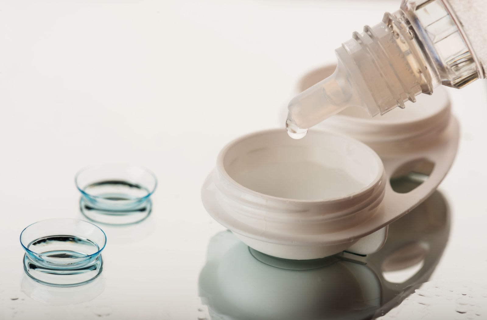 A pair of contact lenses sitting next to a contact lens case and someone pouring contact lens solution into the case