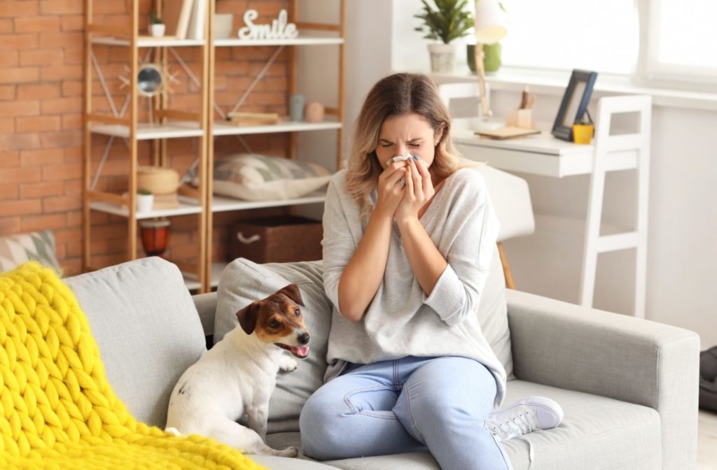 A woman sitting on a couch with her dog and she is blowing her nose with a tissue as she is suffering from allergies
