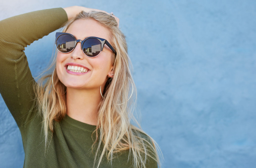 A blonde woman smiling, holding back her hair with her hand, and wearing sunglasses.