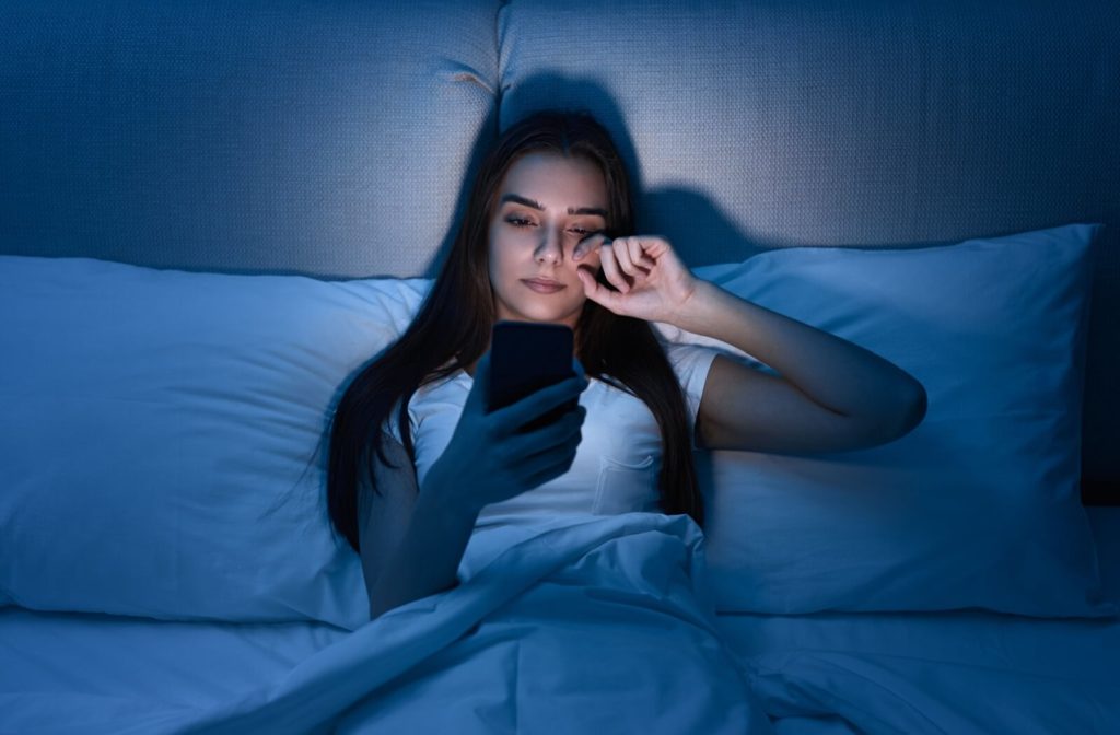 A young woman using her phone in bed while rubbing her dry eyes.