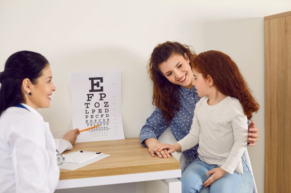 A mom and her daughter attend a child's eye exam, where a friendly eye doctor points to a chart.