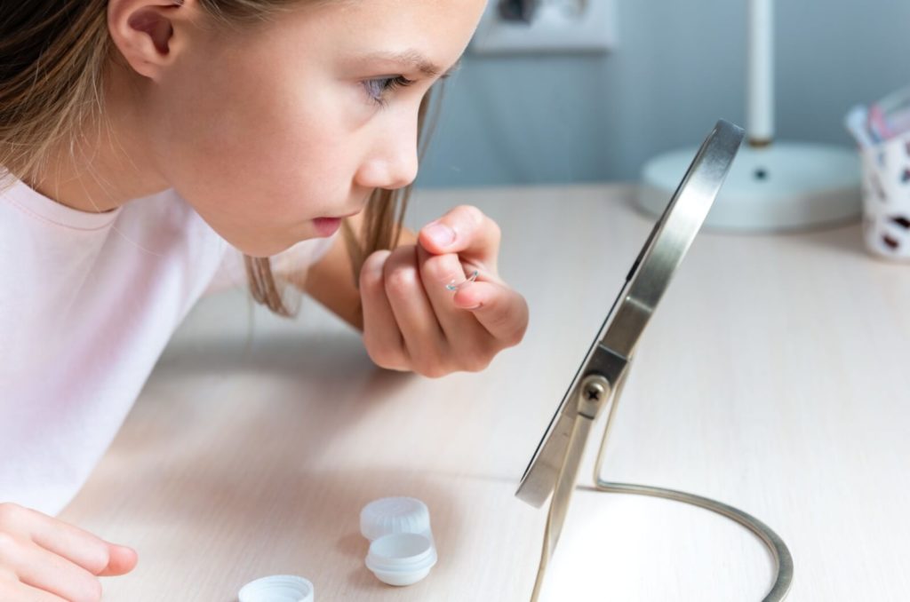 A young girl leans into a mirror to put her contact lenses in.