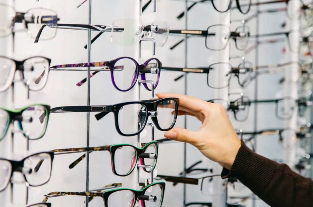 A person's hand reaches for a pair of stylish, vintage-inspired black frames from a wall of glasses at a glasses store.