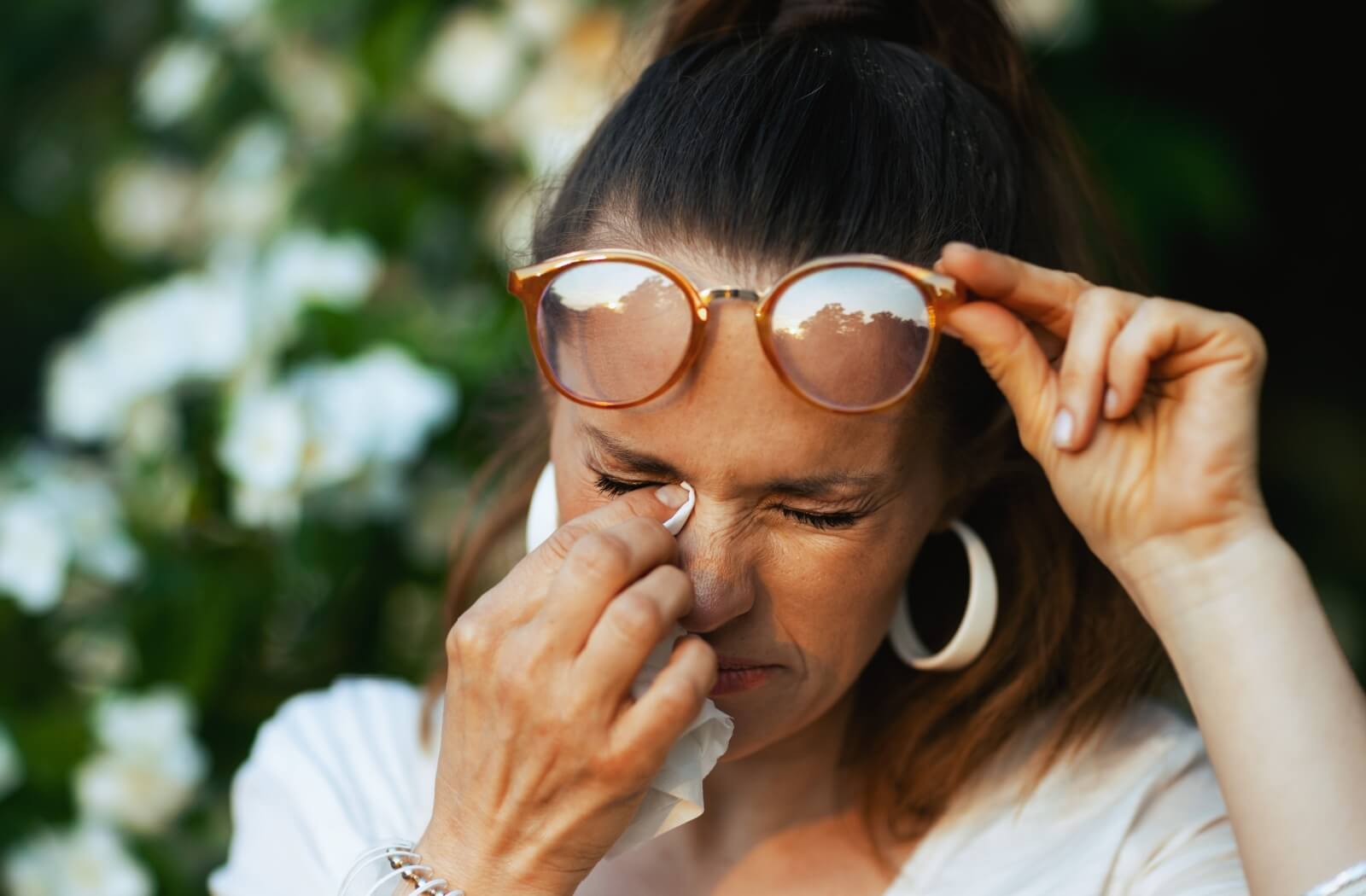 A person lifting their glasses to dab their eyes with a napkin while outdoors due to tearing from dry eyes and allergies.