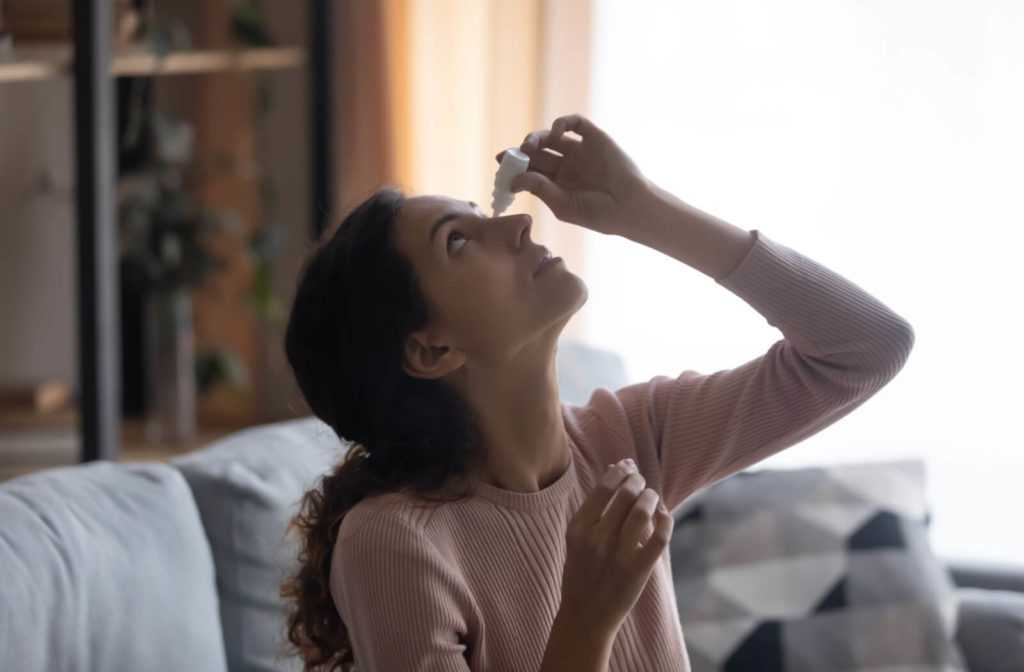 A person sitting on their couch and carefully applying eye drops to their left eye to find relief from allergies and dry eyes.