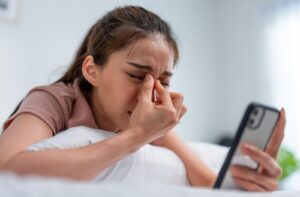 A young adult pinches the bridge of their nose to alleviate dry eye symptoms while on their phone after waking up