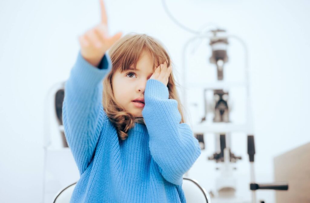 Child in a blue sweater covering one eye during an eye exam, pointing forward while identifying objects on a vision test chart.