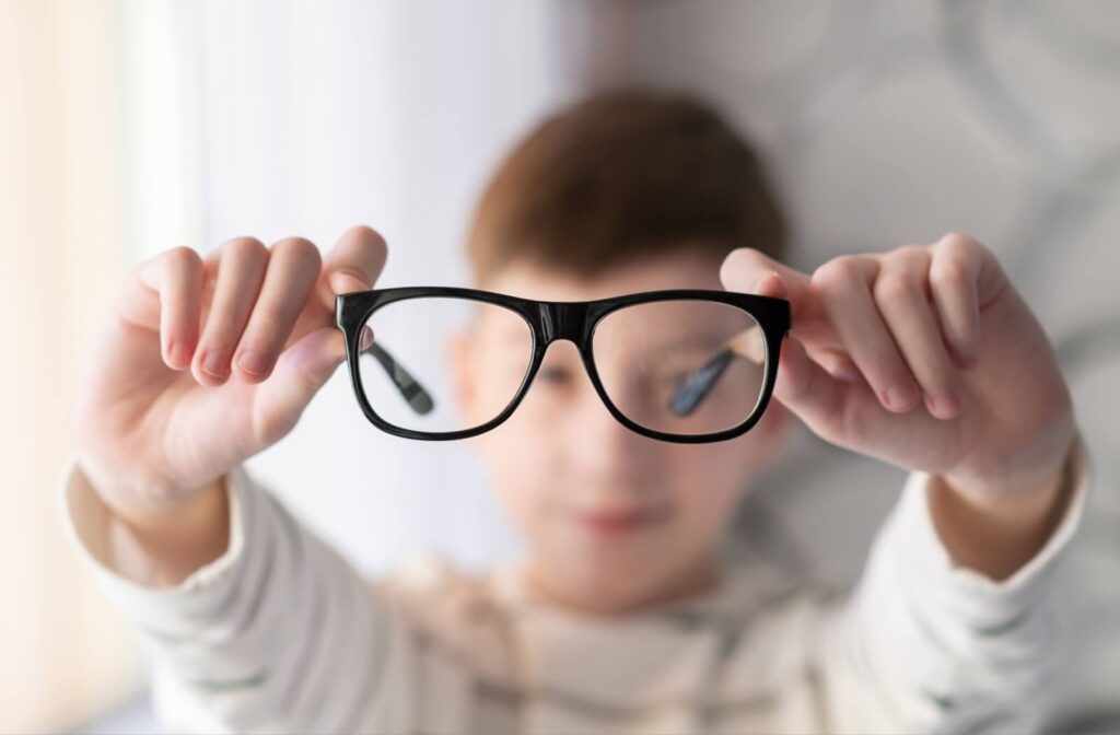  Child holding up a pair of black eyeglasses, with the lenses in focus and the child blurred in the background.