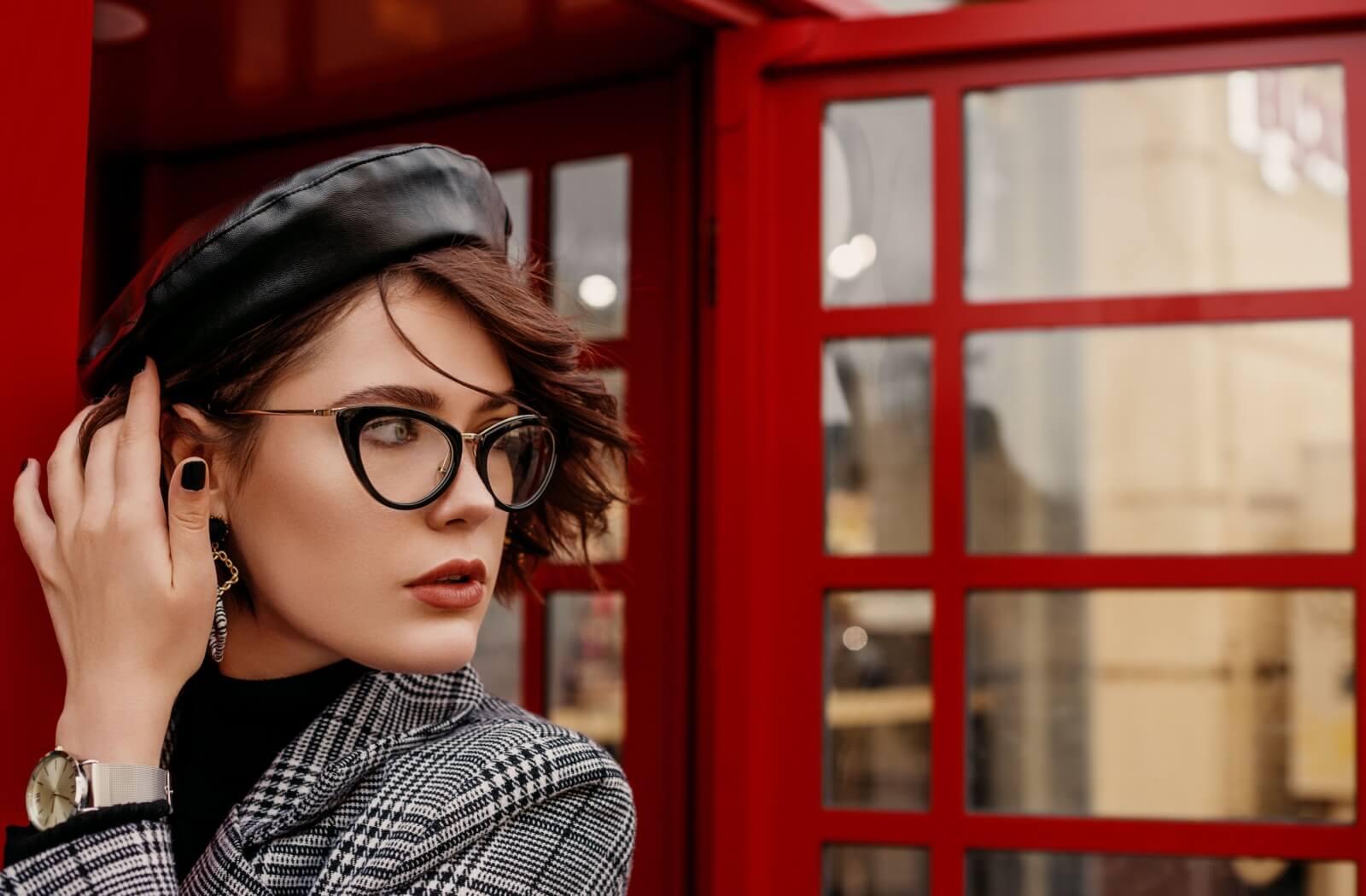 A person wearing black eyeglasses and a beret standing near a red phone booth.