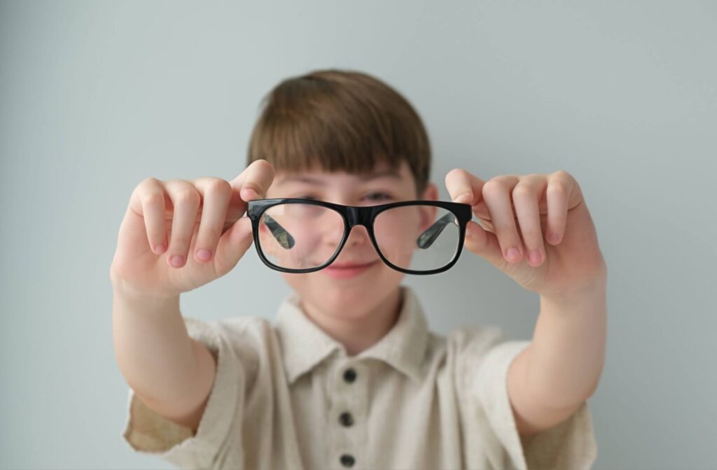 A child holding a pair of black-framed eyeglasses toward the camera with a blurred, smiling face in the background.