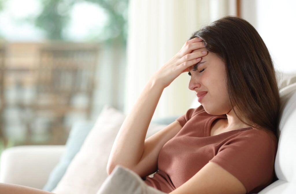 A young woman sitting on a couch, closing her eyes and holding her forehead in pain, illustrating the link between dry eye syndrome and tension headaches.