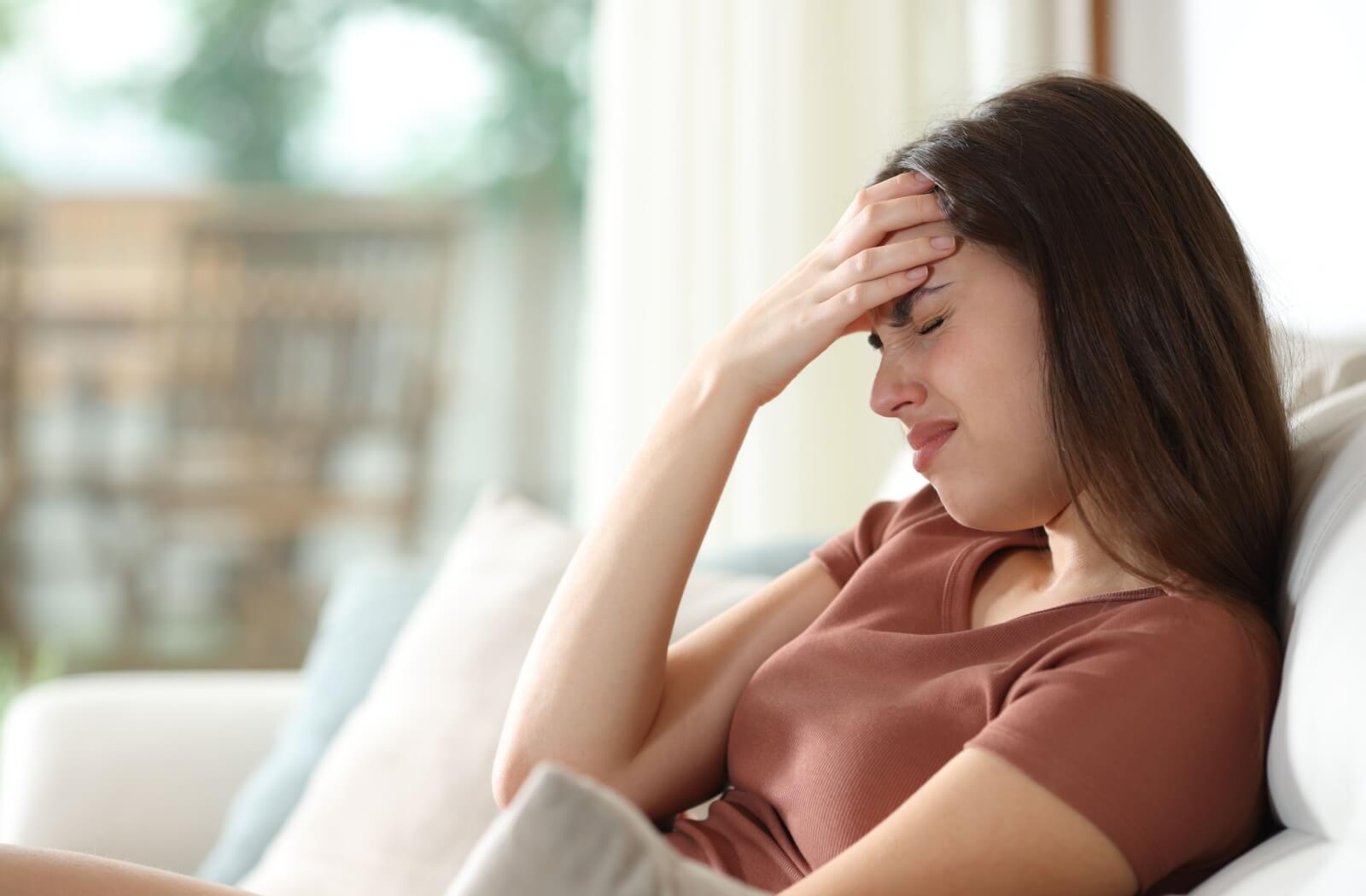 A young woman sitting on a couch, closing her eyes and holding her forehead in pain, illustrating the link between dry eye syndrome and tension headaches.