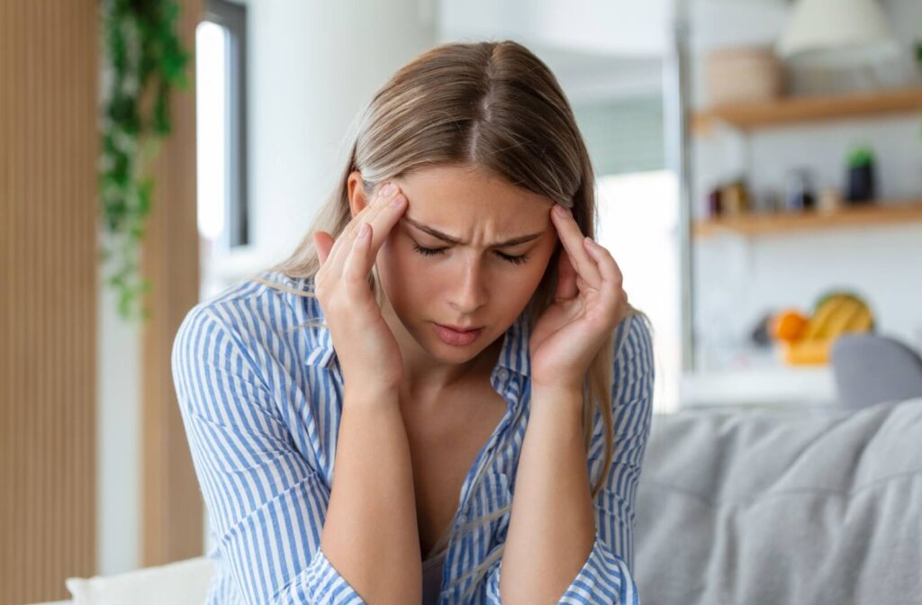 A young woman in a striped shirt massaging her temples with her eyes closed, illustrating a migraine or headache caused by severe dry eye.