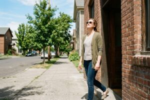 Adult wearing sunglasses leaning against a brick building on a sunny residential sidewalk, looking upward with a relaxed expression.